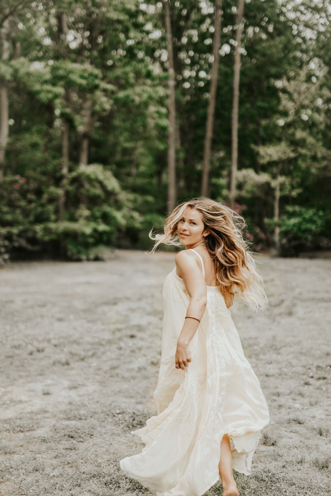 Eine Frau in einem lockeren weißen Kleid läuft zu einem Wald und schaut zurück