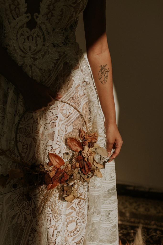 Bildausschnitt von einem Spitzenkleid. Die Frau hält einen Metallring mit Trockenblumen in der Hand.