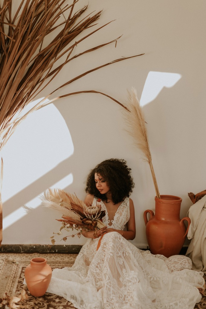 Frau sitzt auf dem Teppich an der Wand und trägt ein Hochzeitskleid aus Spitze, in der Hand hält sich einen Strauß Trockenblumen.