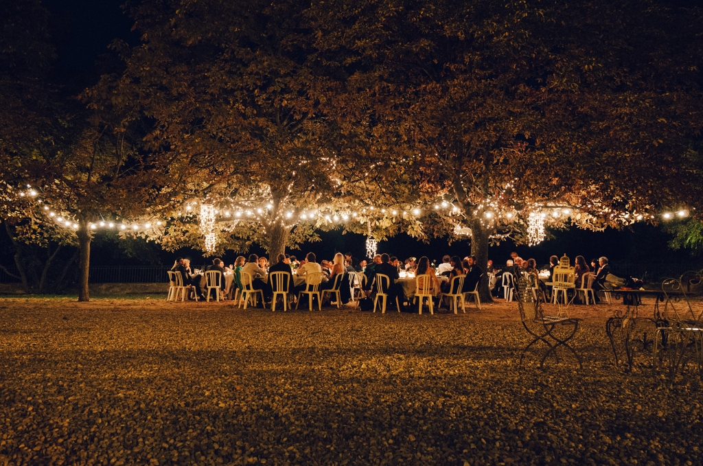 Hochzeitsgesellschaft sitzt unter Bäumen, die mit Lichterketten geschmückt sind